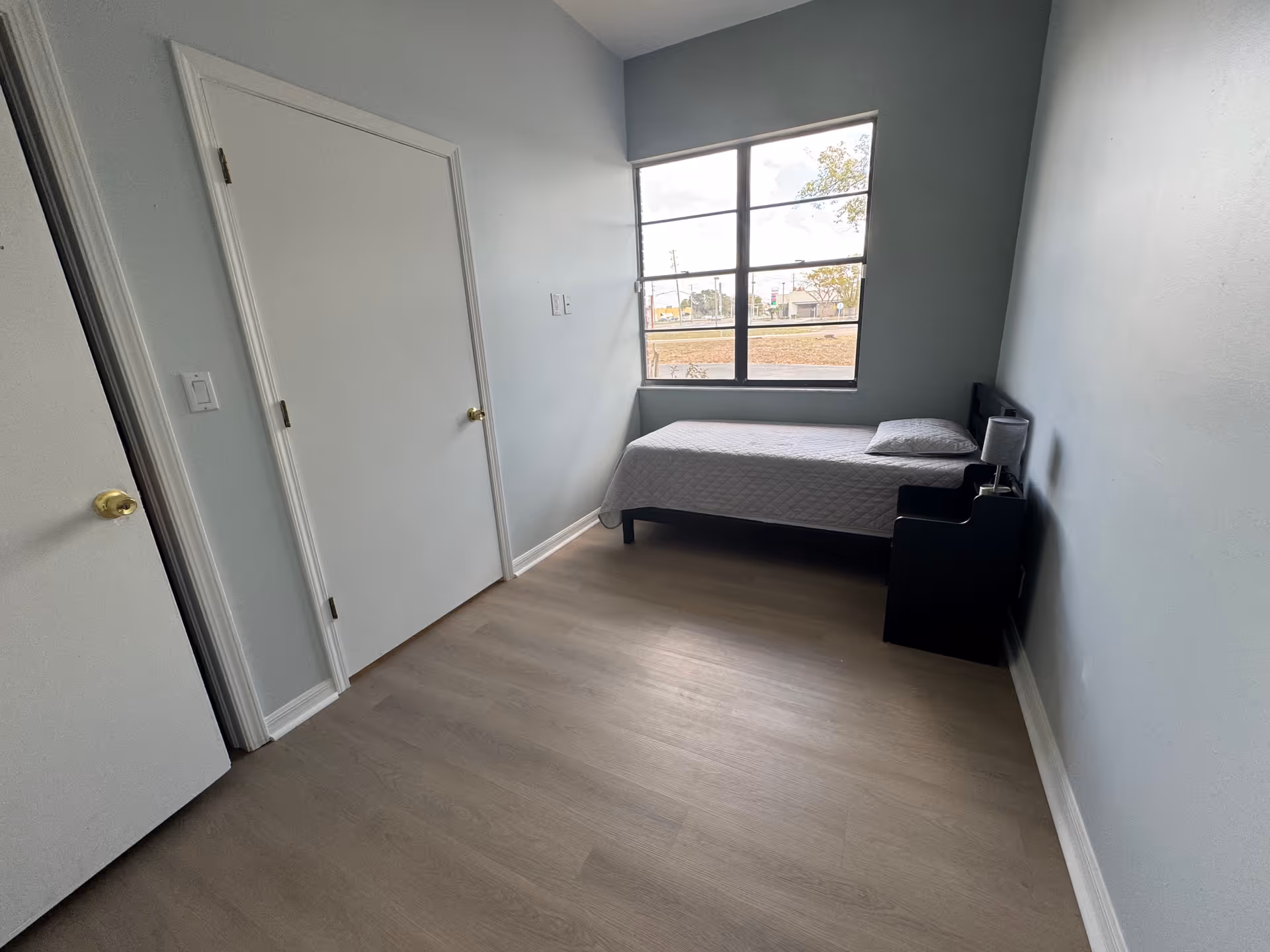A small, simple bedroom with light blue walls and light wood flooring. There is a single bed with a white quilt and pillow positioned against the right wall under a large window. Next to the bed is a small black nightstand with a lamp. Two closed white doors are visible on the left wall.