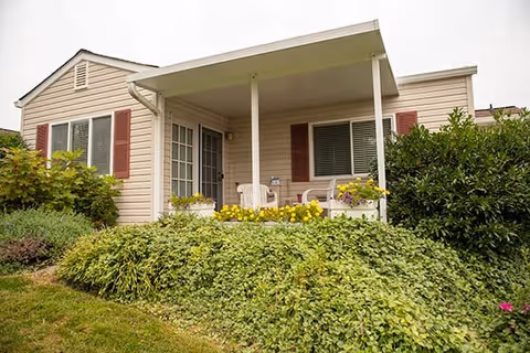 Exterior view of a single-story residential building with beige siding and red shutters. The building features a covered porch with white chairs and a small table, surrounded by green bushes and yellow flowers.