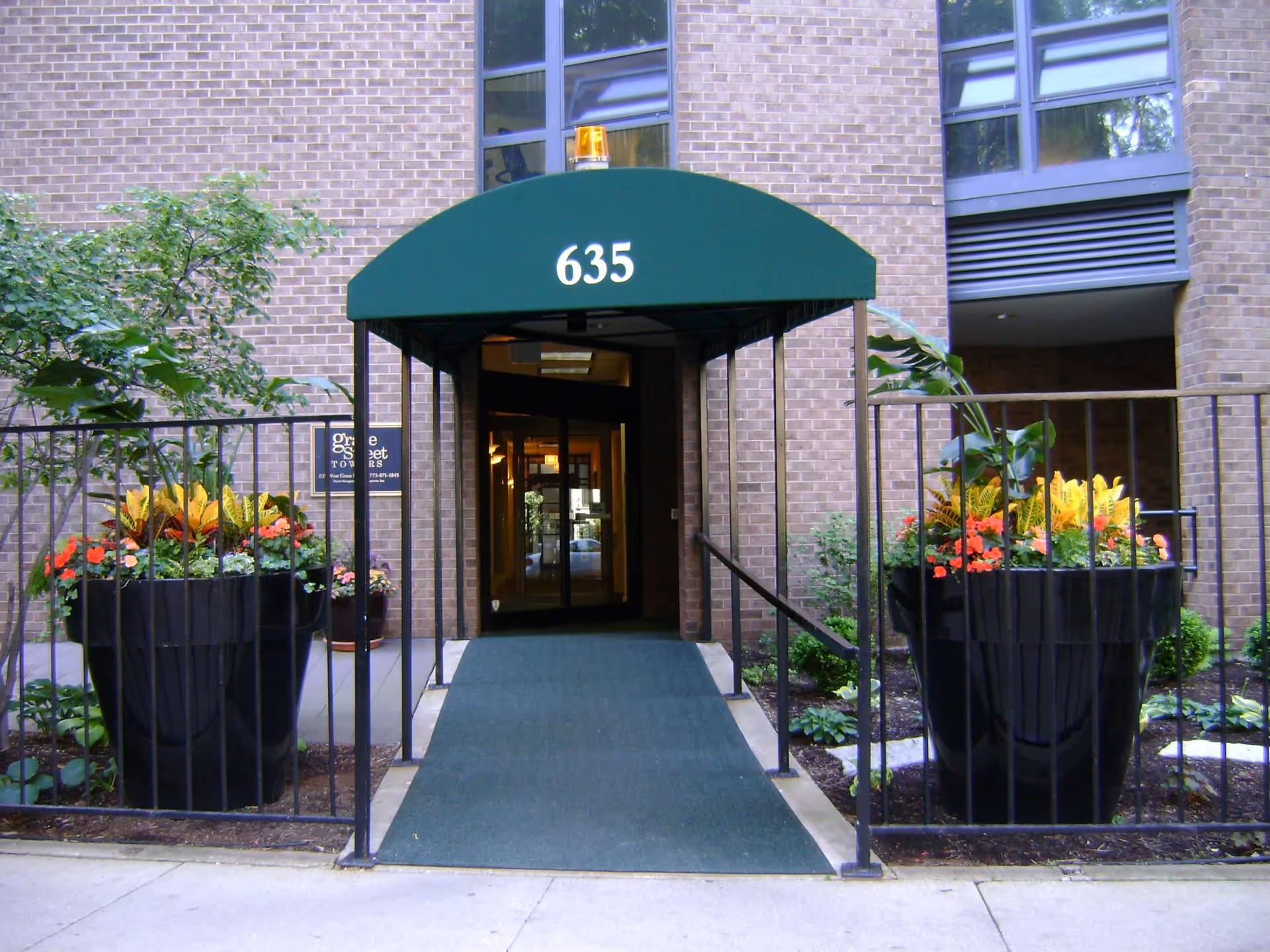 Entrance to a brick building with a green awning displaying the number 635. There is a ramp leading to glass doors, flanked by black railings and large black planters filled with colorful flowers and greenery. A sign on the left side reads 'Grace Street Towers'.