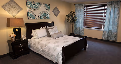 A bedroom with a dark wooden bed frame and white bedding featuring a subtle pattern. There are multiple pillows on the bed. To the left of the bed is a dark wooden nightstand with a lamp, a small clock, and a flower vase. The walls are decorated with three pieces of wall art. A window with blue patterned curtains and closed blinds is on the right side of the room. A potted plant is placed in the corner near the window.