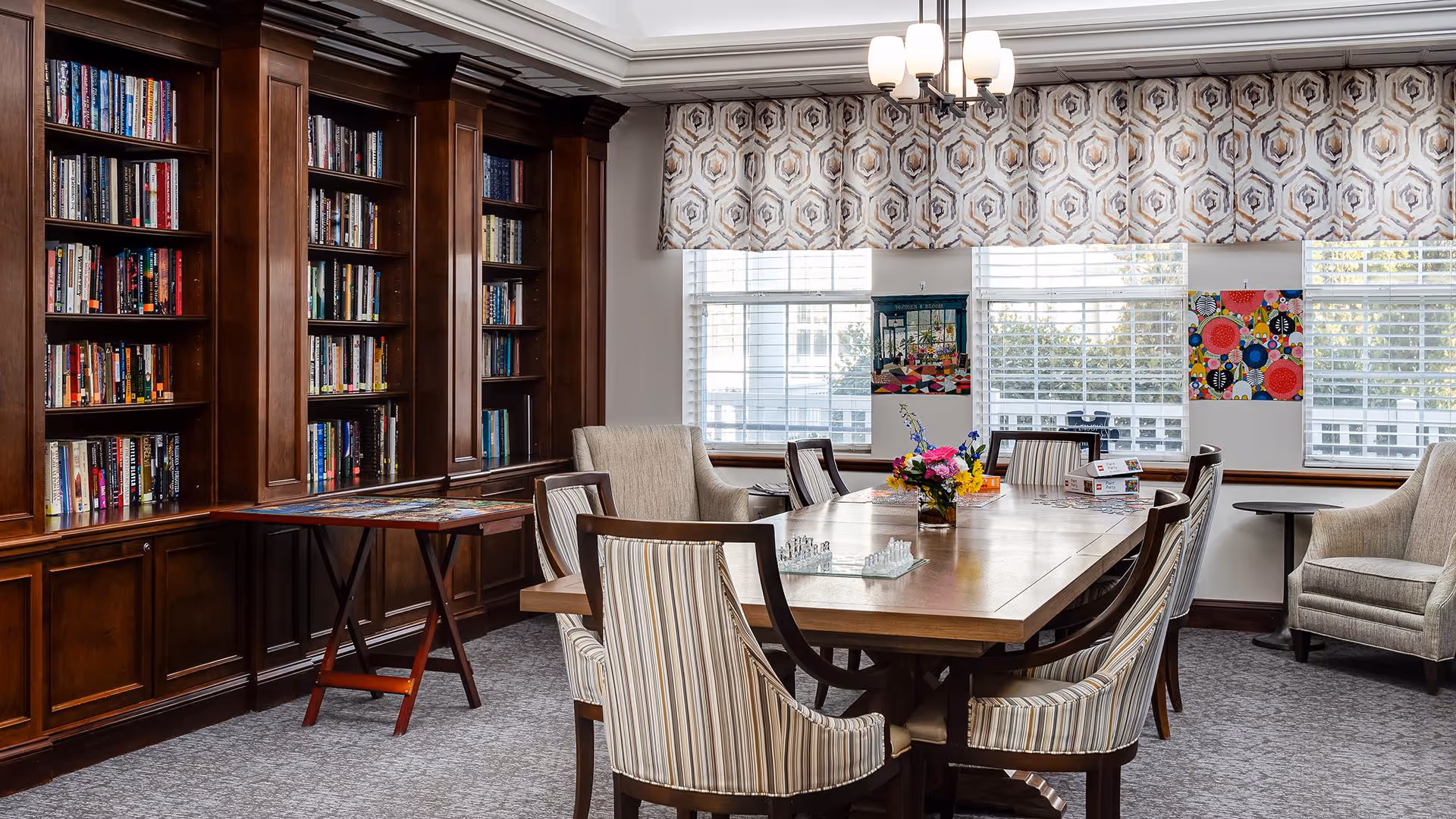A bright communal library/activity room with a long wooden table surrounded by striped chairs, built-in bookshelves, and windows.