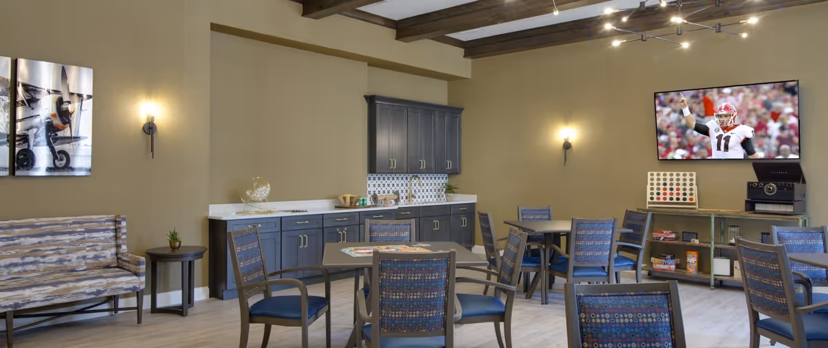 A common area with multiple tables and chairs arranged for socializing or activities. The room features a kitchenette with dark cabinets, a countertop, and a small sink. On the wall, there is a flat-screen TV displaying a football player, and a game board is set up on a console table beneath the TV. The room has beige walls, wood beam ceiling accents, and modern wall sconces providing lighting. A bench and small round table with a plant are also visible.