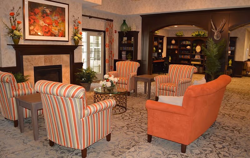 A cozy living room area with four striped armchairs and one orange sofa arranged around a glass coffee table with a floral centerpiece. The room features a fireplace with a floral painting above it, potted plants, patterned carpet, and a bookshelf in the background with decorative items and a mounted deer head on the wall.