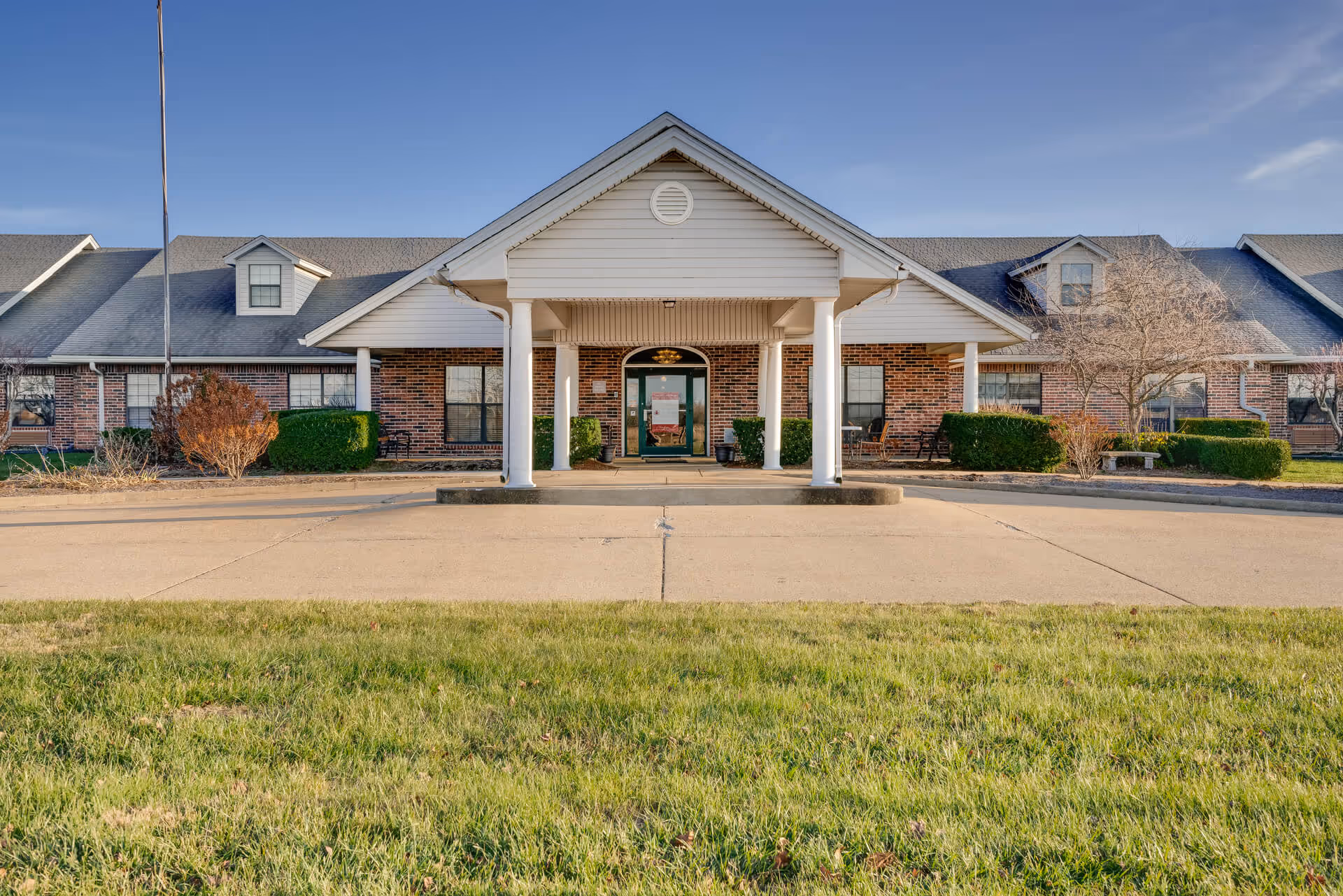 Front exterior view of a single-story brick building with a covered entrance supported by white columns. The building has multiple windows, a gray shingled roof with dormer windows, and a concrete driveway leading up to the entrance. There is a grassy lawn in the foreground and a clear blue sky above.
