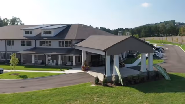 Exterior view of a senior living facility building with a covered entrance, multiple windows, and a parking lot with cars. The building is surrounded by green grass and trees in the background under a clear sky.