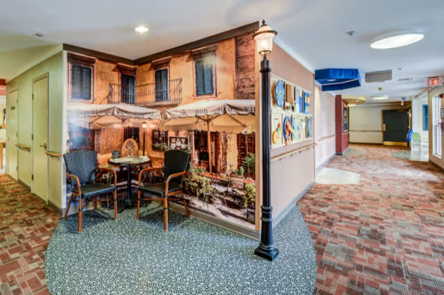 A decorated interior hallway in a memory care facility featuring a faux outdoor café mural with a small table, two chairs, and a lamppost.