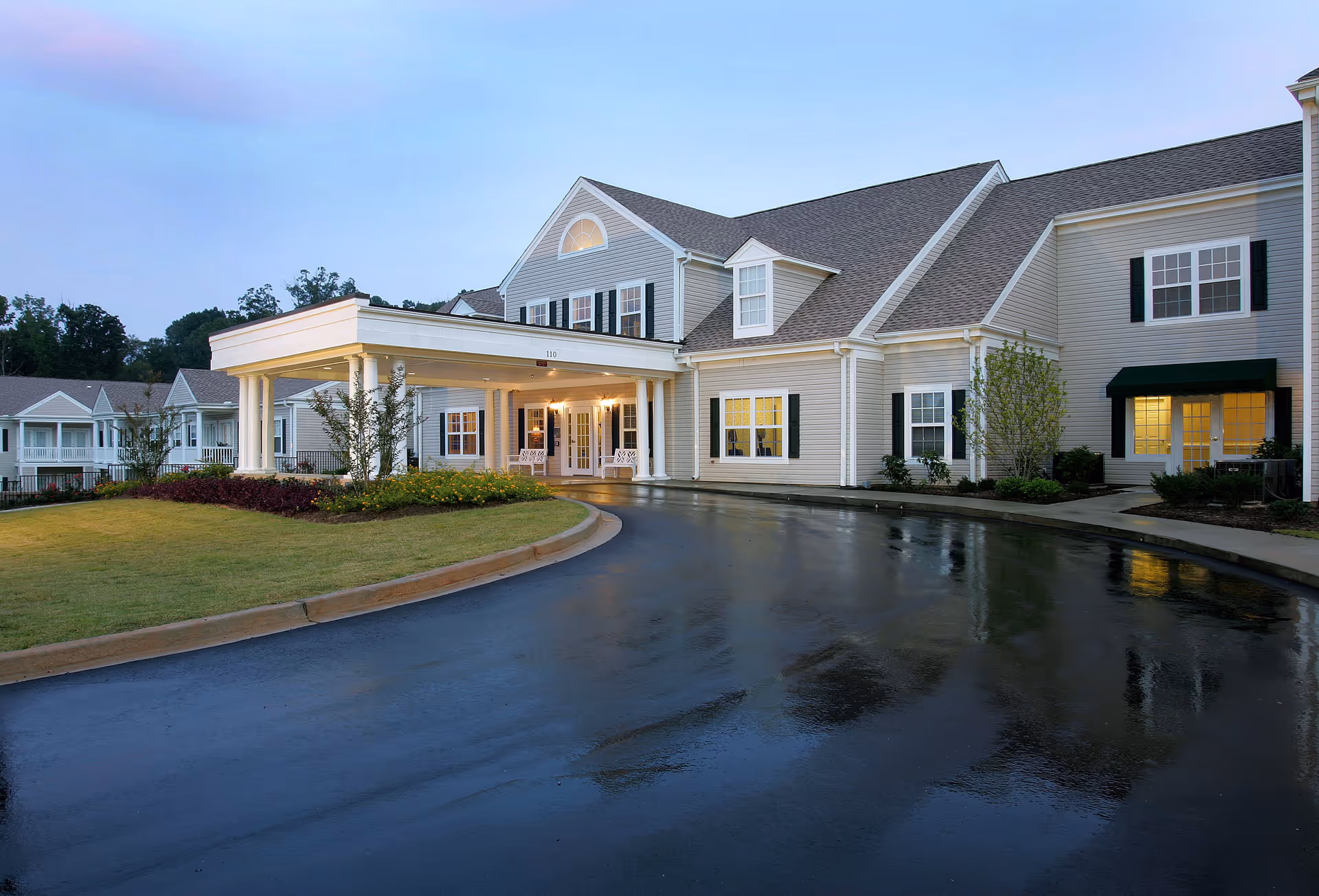 Exterior view of The Willows of Easley senior living facility at dusk, showing a large, two-story building with white siding, black shutters, and a covered entrance with columns. The driveway is wet, reflecting the building's lights, and there is a well-maintained lawn and landscaping around the entrance.