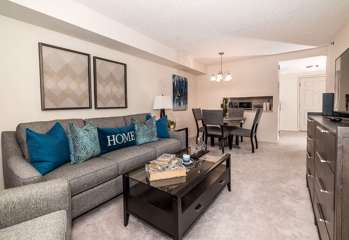 A cozy living room with a gray sofa adorned with blue and patterned throw pillows, including one with the word 'HOME'. A glass-top coffee table with decorative books and a candle sits in front of the sofa. To the right, there is a dark wood dresser with a flat-screen TV on top. In the background, a dining area with a round table and four chairs is visible, along with a hanging light fixture and a small pass-through window to the kitchen. The room is carpeted and decorated with framed artwork on the walls.