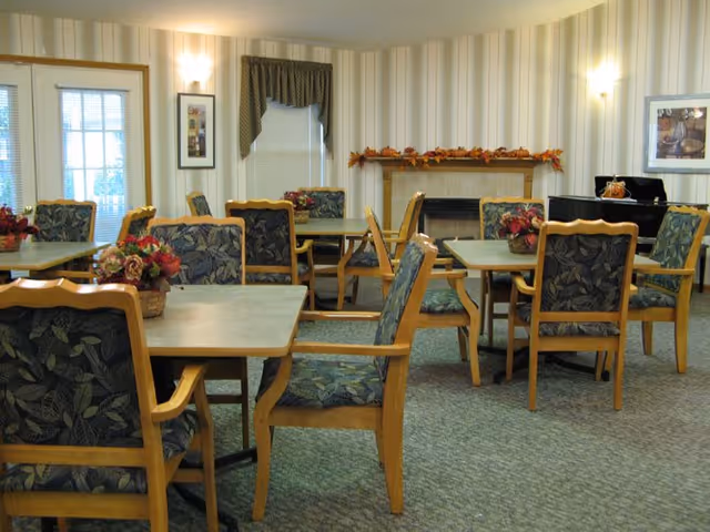A cozy dining room with several tables and wooden chairs upholstered with leafy patterned fabric. Each table has a small floral centerpiece. The room features a fireplace decorated with autumn leaves, a piano in the corner, and windows with curtains allowing natural light to enter.