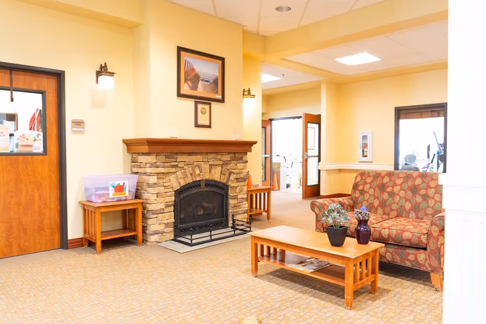 A cozy interior common area with a stone fireplace, a patterned sofa, a wooden coffee table with two potted plants, and wooden side tables. The walls are painted light yellow, and there are framed pictures above the fireplace. Doors and windows are visible in the background.