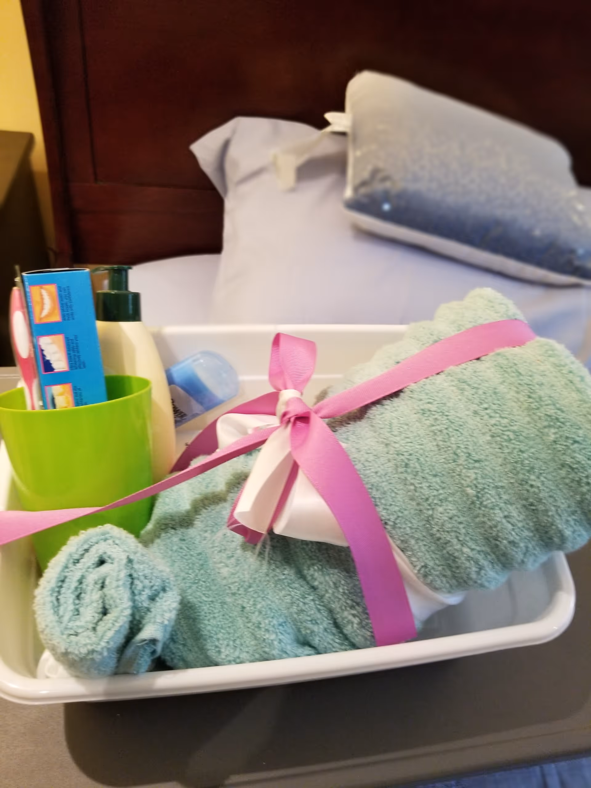 A white tray on a bed with a rolled green towel tied with a pink ribbon, a green cup holding a toothbrush and toothpaste, and bottles of lotion and deodorant, with pillows and a wooden headboard in the background.