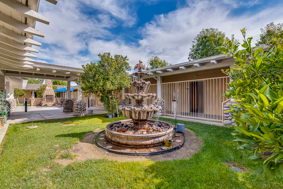 Outdoor courtyard area at Sunny Rose Senior Living featuring a multi-tiered stone water fountain surrounded by green grass and trees, with covered walkways and building entrances in the background under a partly cloudy sky.