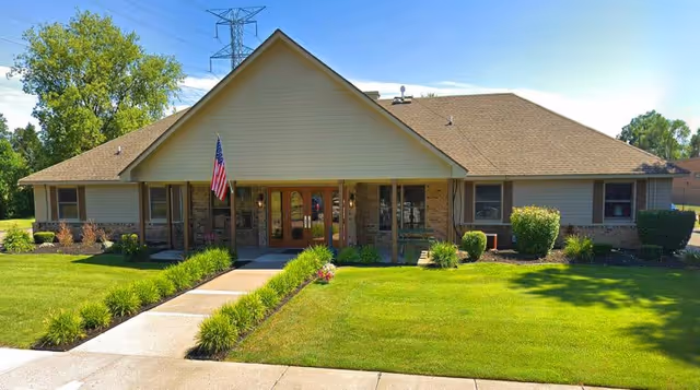 Single-story building with a gable roof and beige siding, featuring a central entrance with double wooden doors and an American flag mounted on a pole near the entrance. The building is surrounded by a well-maintained lawn with shrubs and small plants along the walkway leading to the entrance.