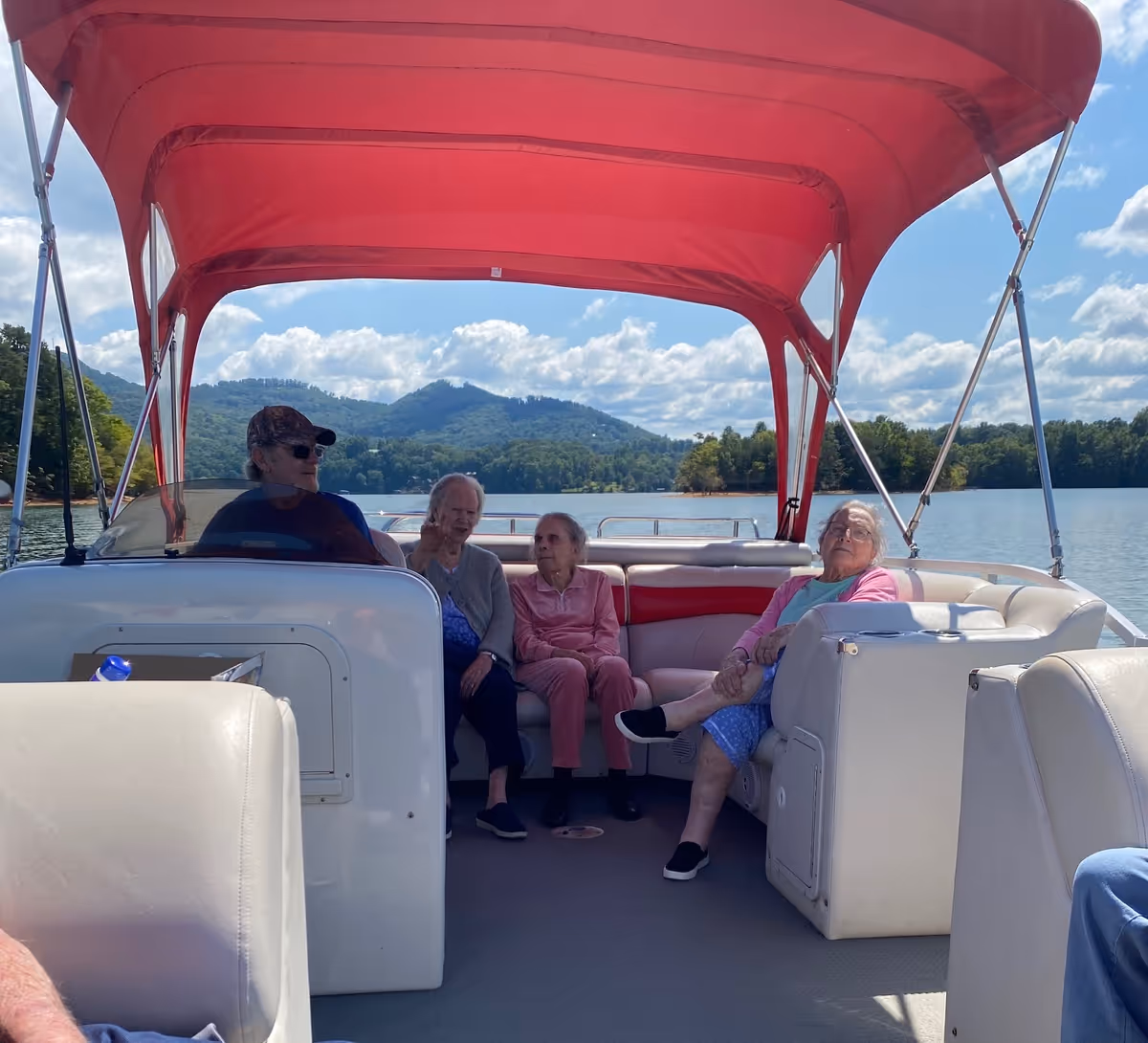 Four elderly people sitting on a boat with a red canopy, surrounded by water and scenic mountains in the background under a partly cloudy sky.