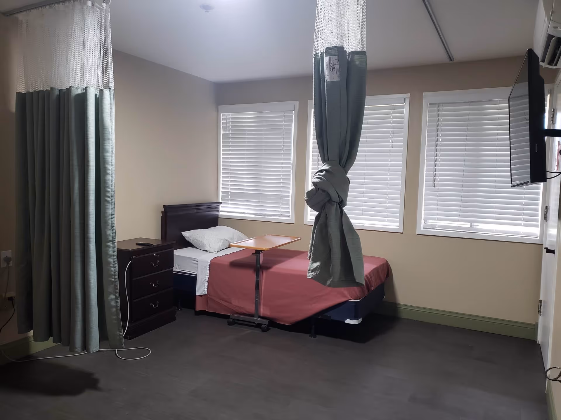 A simple bedroom in a senior living facility with a single bed covered with a red blanket and a white pillow. There is a small bedside table with drawers next to the bed, and a rolling overbed table positioned on the bed. Three windows with white blinds are on the wall behind the bed. A green privacy curtain is partially drawn and tied up near the bed. A flat-screen TV is mounted on the wall to the right.