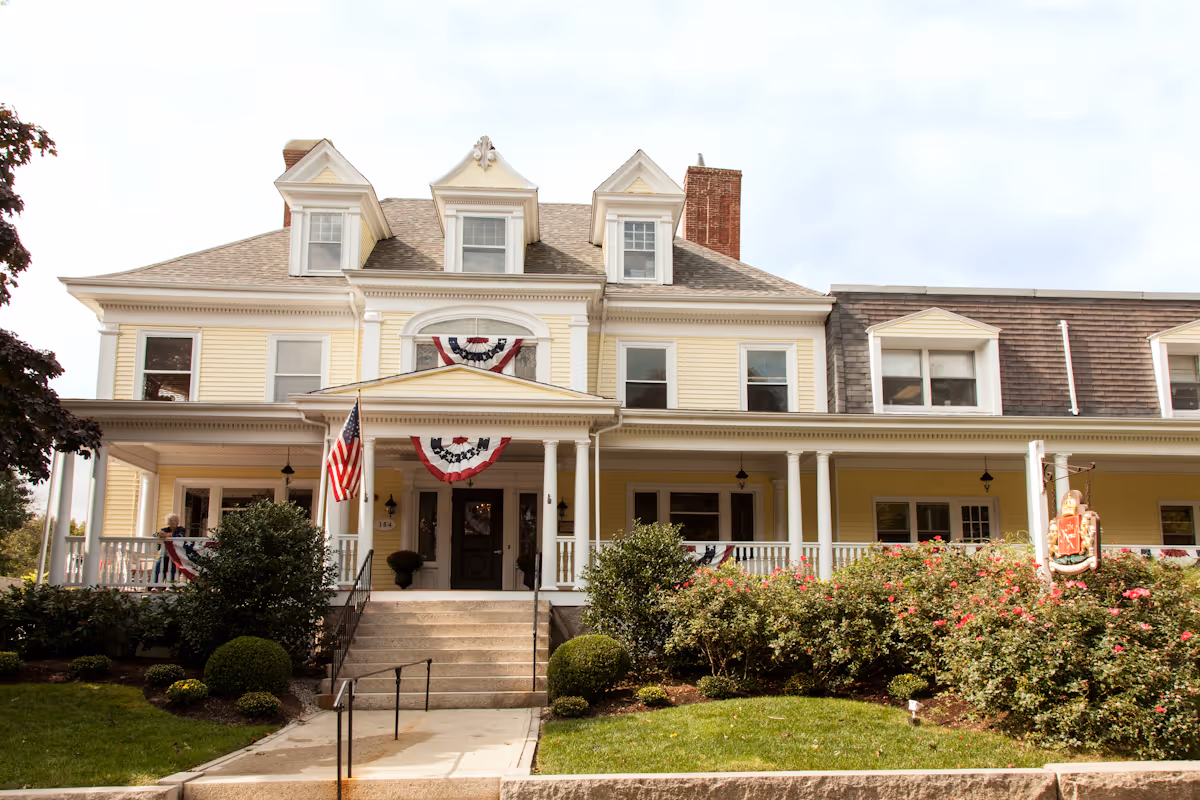 Front facade of a large yellow Victorian-style nursing center with a wraparound porch, American flag and decorative bunting, and landscaped steps leading to the entrance.