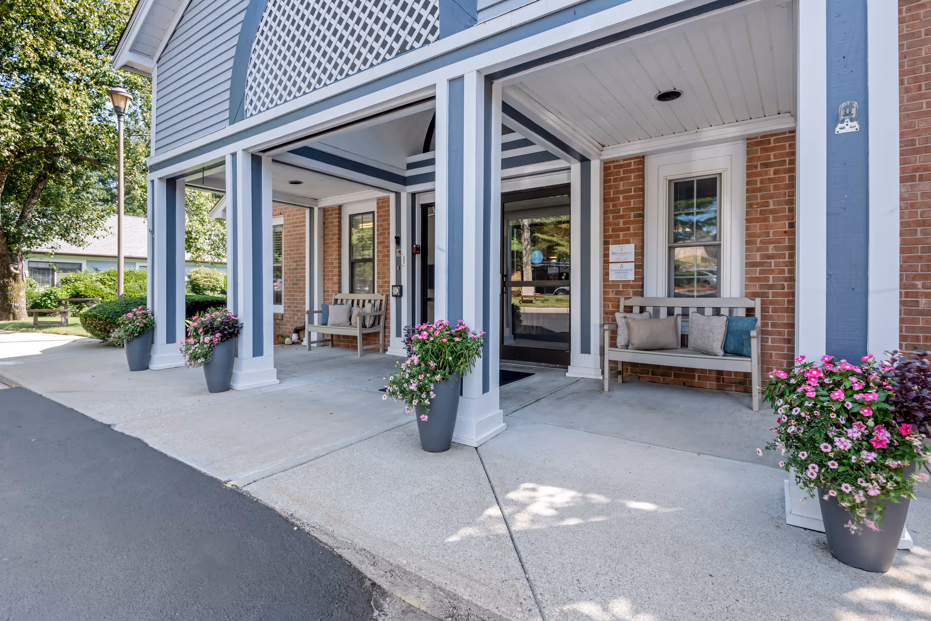 Entrance area of a senior living facility with a covered porch supported by blue and white columns. There are two wooden benches with cushions placed against a brick wall, and large flower pots with pink and white flowers on either side of the entrance. Trees and greenery are visible in the background.