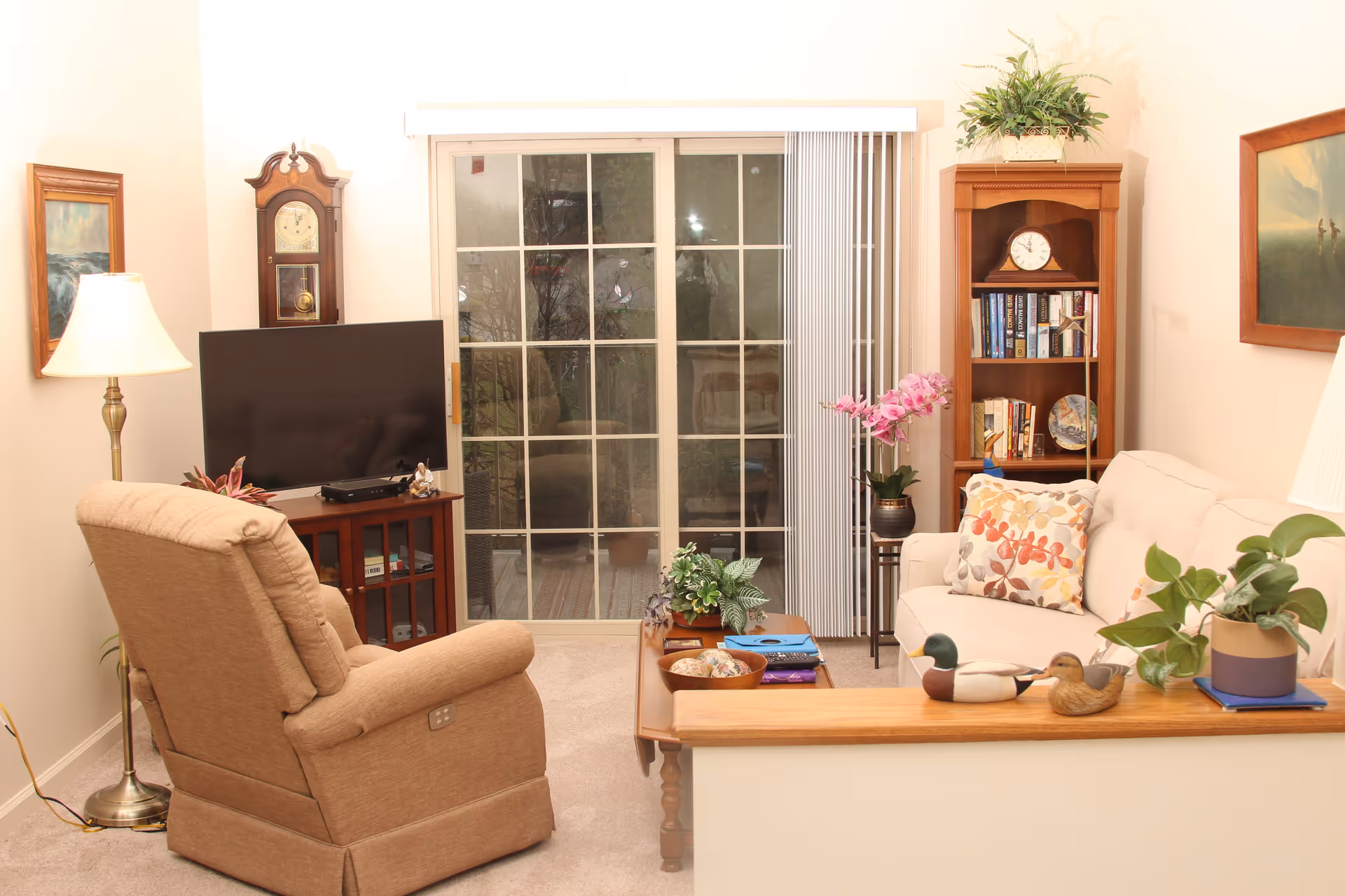 A cozy living room with a beige recliner chair, a white sofa with floral cushions, a wooden coffee table with plants and decorative items, a wooden bookshelf filled with books and a clock, a flat-screen TV on a wooden stand, a grandfather clock, two framed paintings on the walls, a floor lamp, and a sliding glass door with vertical blinds leading outside.