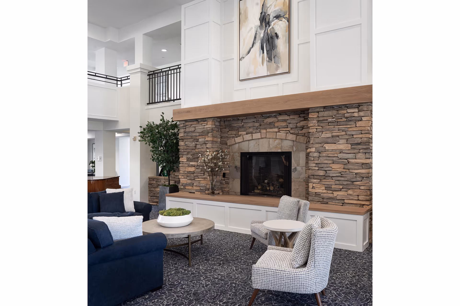 A cozy living room area featuring a stone fireplace with a wooden mantel, a large abstract painting above it, a navy blue sofa with white pillows, two patterned armchairs with cushions, a round coffee table with a white planter, and a small side table. The room has a patterned carpet and white walls with decorative paneling.
