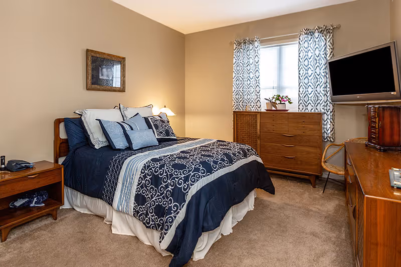 A cozy bedroom featuring a bed with a navy blue and white patterned comforter and multiple pillows. The room has beige walls and carpeted floor. There is a wooden nightstand with a telephone and lamp on one side of the bed. Across from the bed, there is a wooden dresser with a flat-screen TV on top and a wooden chest of drawers beneath a window with patterned curtains. A small chair is placed next to the dresser.