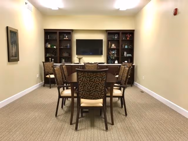 A small meeting or dining room with a rectangular wooden table surrounded by eight cushioned chairs with patterned backs. At the far end of the room, there are two dark wooden bookshelves flanking a mounted flat-screen TV. The shelves contain various decorative items. The walls are painted light yellow, and the floor is carpeted in a neutral tone. A framed picture hangs on the left wall.