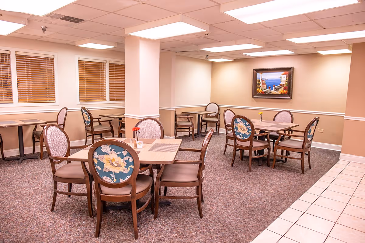 Dining room with several wooden tables and floral-upholstered chairs, a painting on the wall, and windows with blinds.