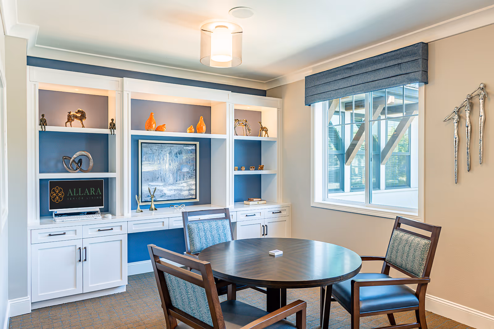 A bright and modern room in Allara Senior Living featuring a round wooden table with four chairs, a built-in white shelving unit with decorative items and a computer displaying the Allara Senior Living logo, a large window with a blue valance, and a wall decoration of three metallic human figures.