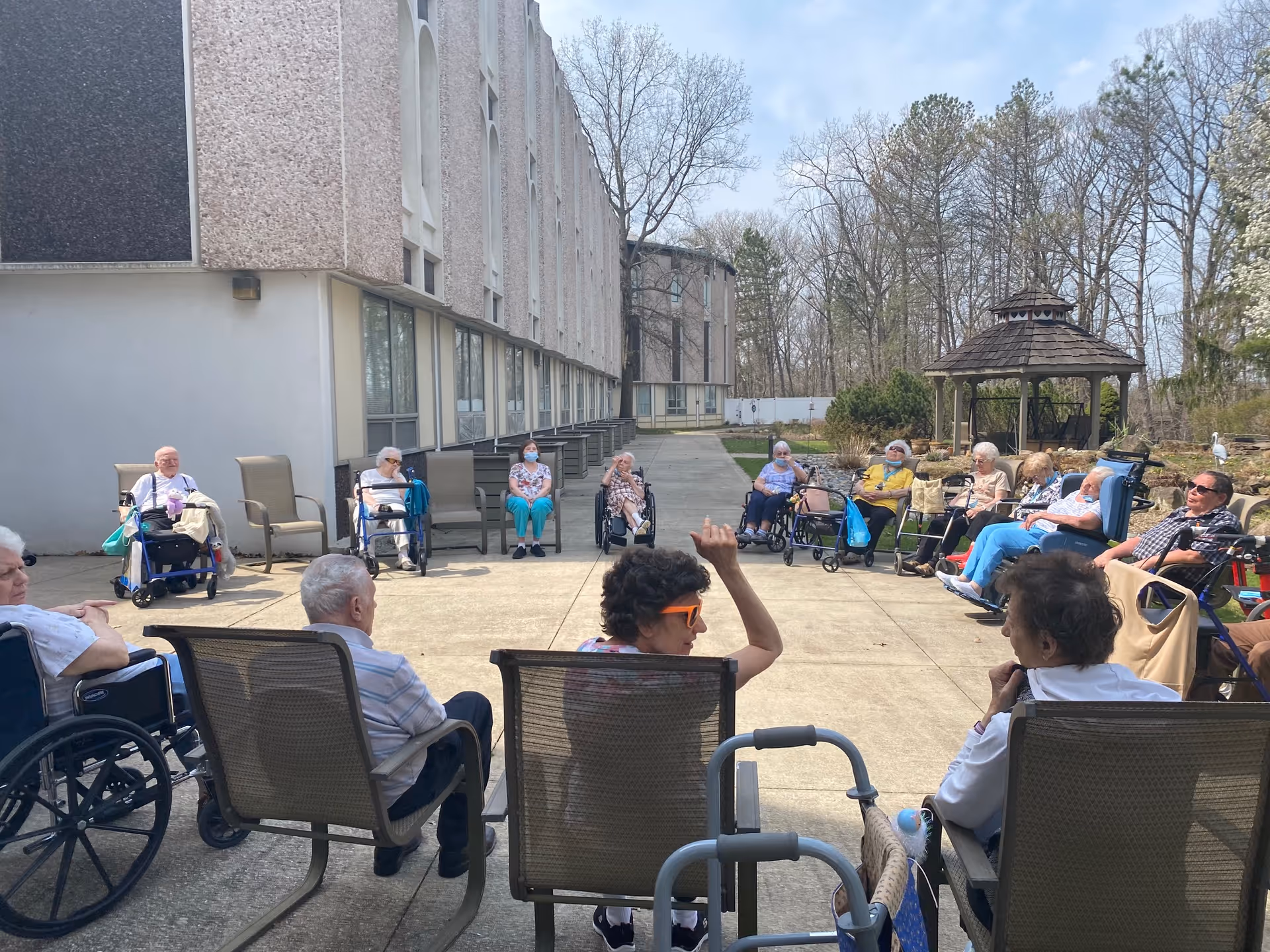 A group of elderly people sitting in a large outdoor courtyard area of a senior living facility. They are seated in a circle on chairs and wheelchairs, some engaging in conversation. The building is visible on the left side, and there is a gazebo and trees in the background under a clear sky.