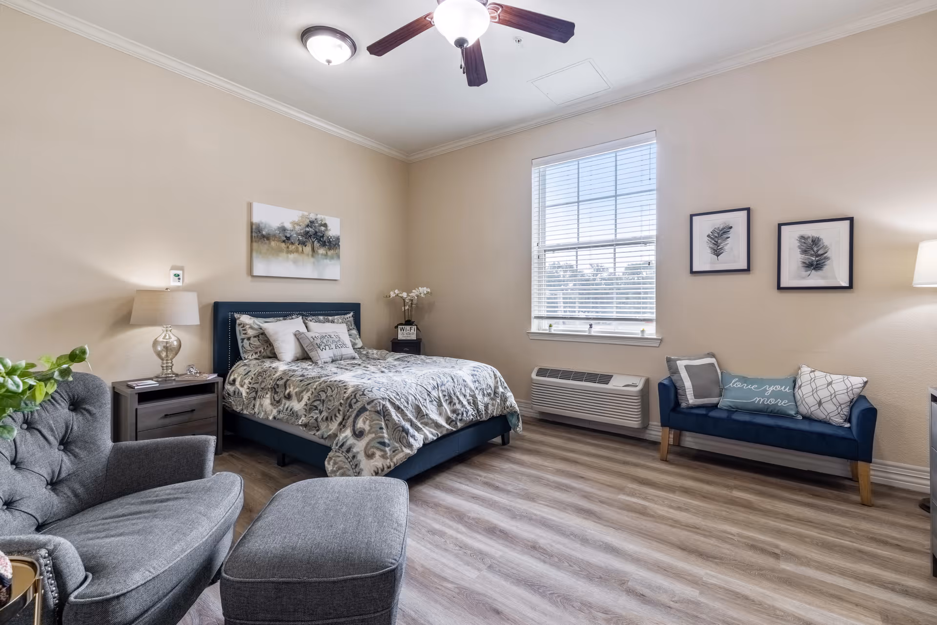 Bright, furnished bedroom with a bed, nightstand, armchair and ottoman, small sofa, and a window letting in natural light.