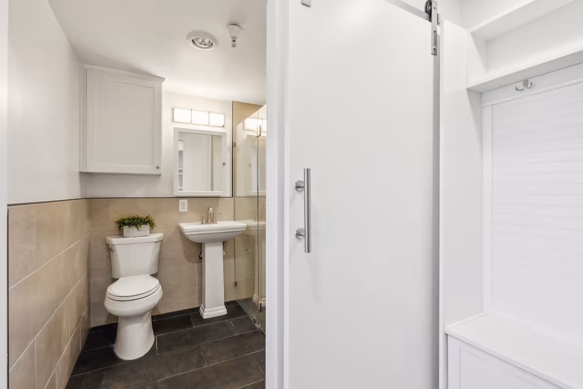 A clean and modern bathroom featuring a white toilet with a small green plant on top, a white pedestal sink with a rectangular mirror above it, beige tiled walls halfway up, and dark floor tiles. To the right, there is a white sliding door partially open revealing a white built-in bench with hooks above it.