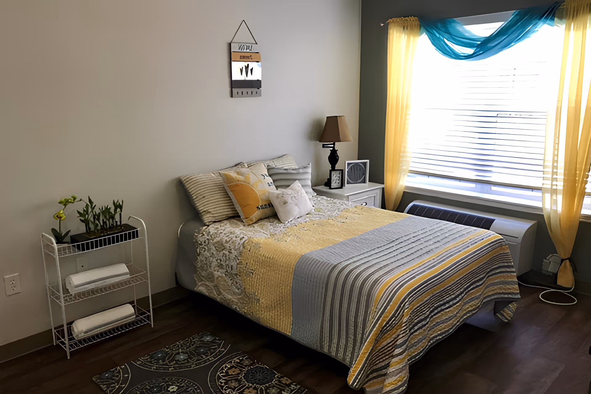 A cozy bedroom with a bed covered in a yellow, gray, and white striped quilt. The bed has several pillows, including one with a sun design. Next to the bed is a white nightstand with a lamp and framed pictures. A window with yellow curtains and a blue sheer swag lets in natural light. A small white metal shelf with potted plants and folded towels is against the wall. The floor is wooden with a small patterned rug near the bed.