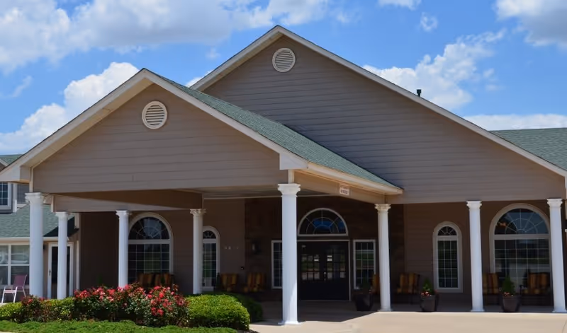 Front exterior view of a senior living facility building with a covered entrance supported by white columns, large arched windows, green roof, and landscaping with bushes and flowers under a partly cloudy sky.
