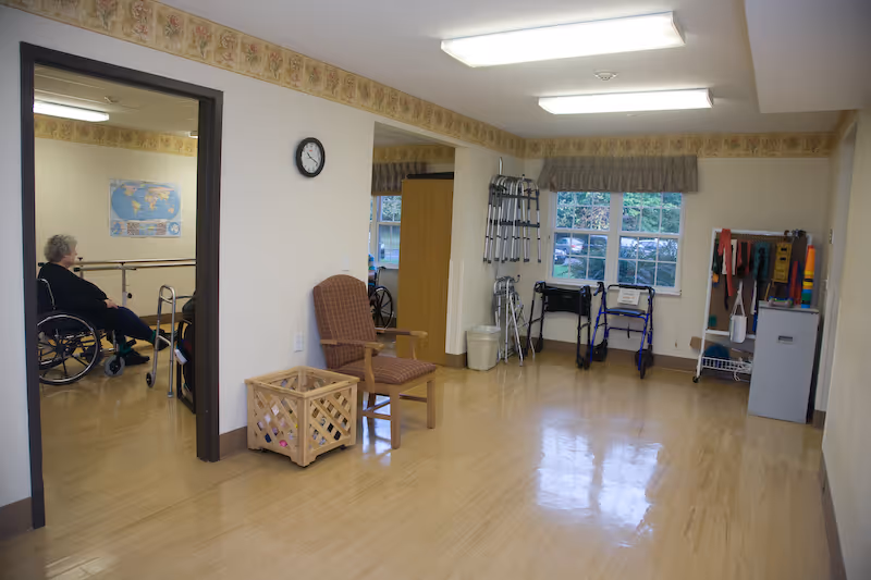 Bright interior therapy/rehab room with chairs, walkers and other mobility aids, and a person in a wheelchair visible through a doorway.