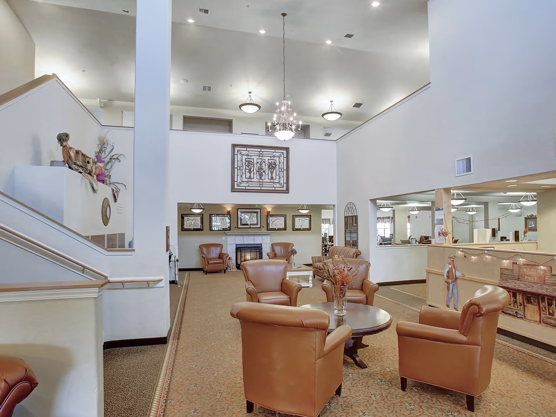 A spacious and well-lit senior living facility common area with high ceilings, beige walls, and carpeted floors. The room features a seating arrangement with four brown leather armchairs around a round wooden table with a vase of flowers. In the background, there is a fireplace flanked by two more armchairs and a couch, with framed artwork above the fireplace. A staircase with wooden handrails is visible on the left side, and a chandelier hangs from the ceiling. The area has a welcoming and comfortable atmosphere.