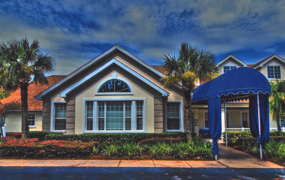 Exterior front view of a residential building with a peaked roof, large windows, palm trees, and a blue canopy covering the entrance walkway. The sky is partly cloudy.