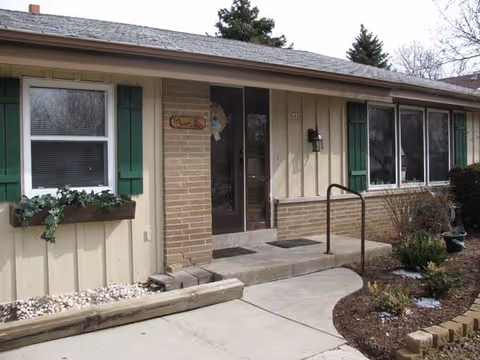 Exterior view of a single-story building entrance with a concrete walkway, green window shutters, a small garden area with plants, and a handrail leading to a glass door.