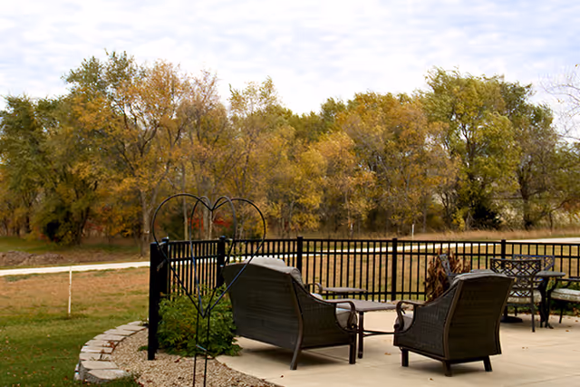 Outdoor seating area with wicker chairs beside a fence amid fall-colored trees.