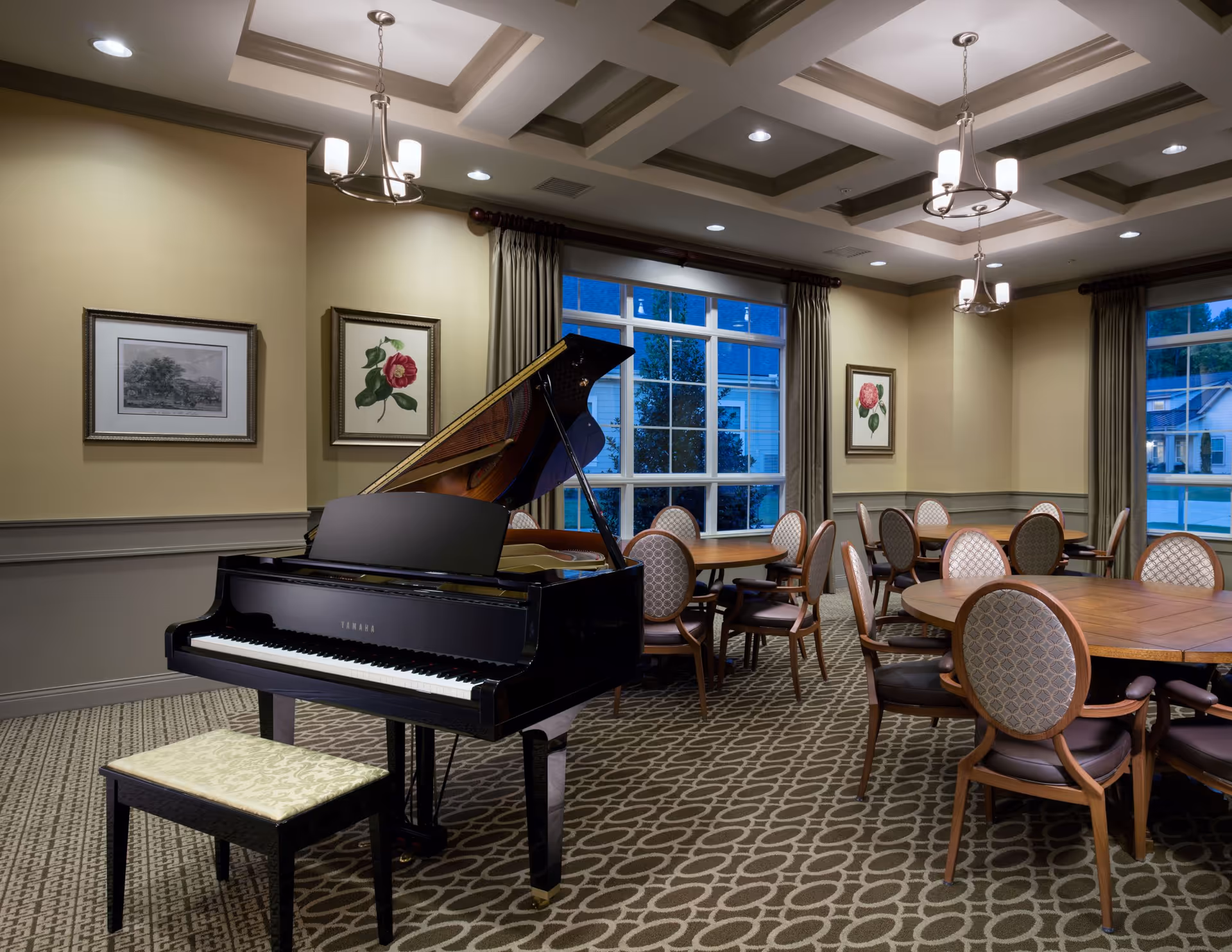 A well-lit communal room with a grand piano, round dining tables and upholstered chairs near large windows.