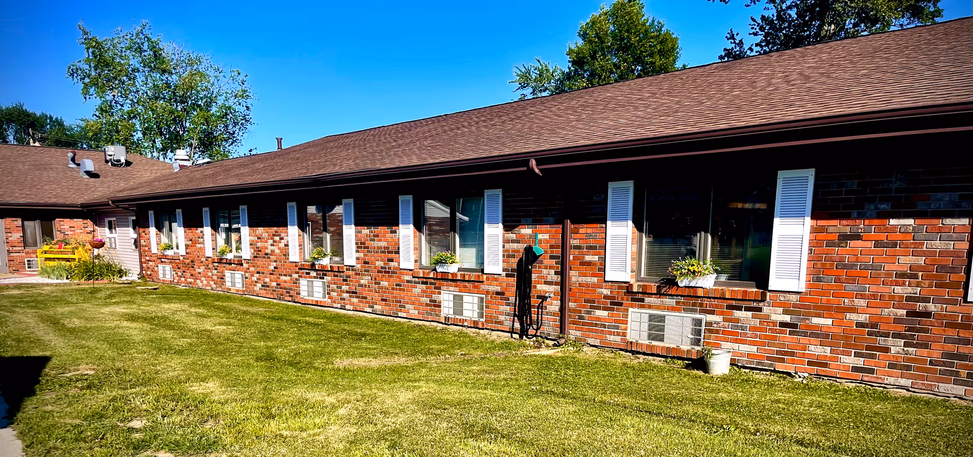 Exterior view of a single-story brick building with white window shutters and flower boxes under the windows. The building has a brown shingled roof and is surrounded by a well-maintained grassy lawn under a clear blue sky.