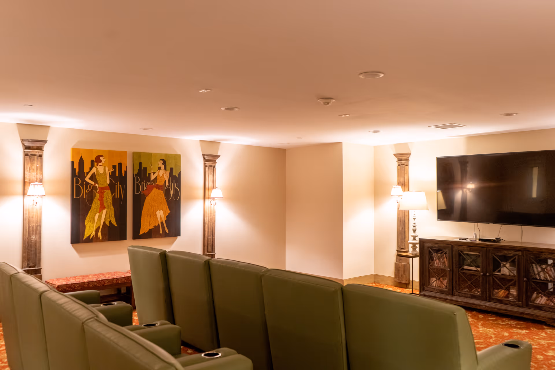 A cozy entertainment room with a row of green recliner chairs facing a large flat-screen TV mounted on the wall. The room features warm lighting from wall sconces and a table lamp, decorative wooden columns, and two colorful paintings of women with the text 'Big City Bright Lights' on the wall.