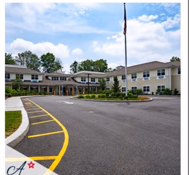 Front exterior view of a two-story assisted living facility with a circular driveway, landscaped greenery, and an American flag on a flagpole in the center of the driveway. The building has multiple windows and a covered entrance.