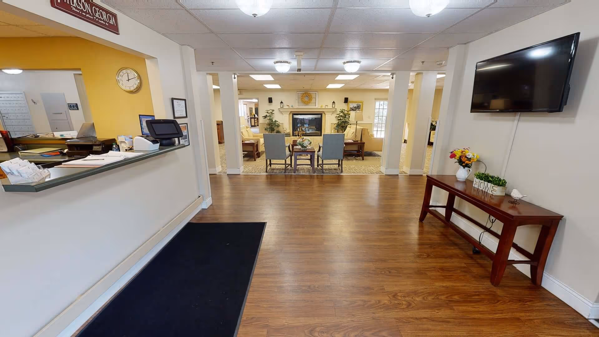 Interior view of a senior living facility lobby with a reception desk on the left, a seating area with chairs and sofas around a fireplace in the background, a wall-mounted TV on the right, and a wooden table with decorative items beneath the TV.