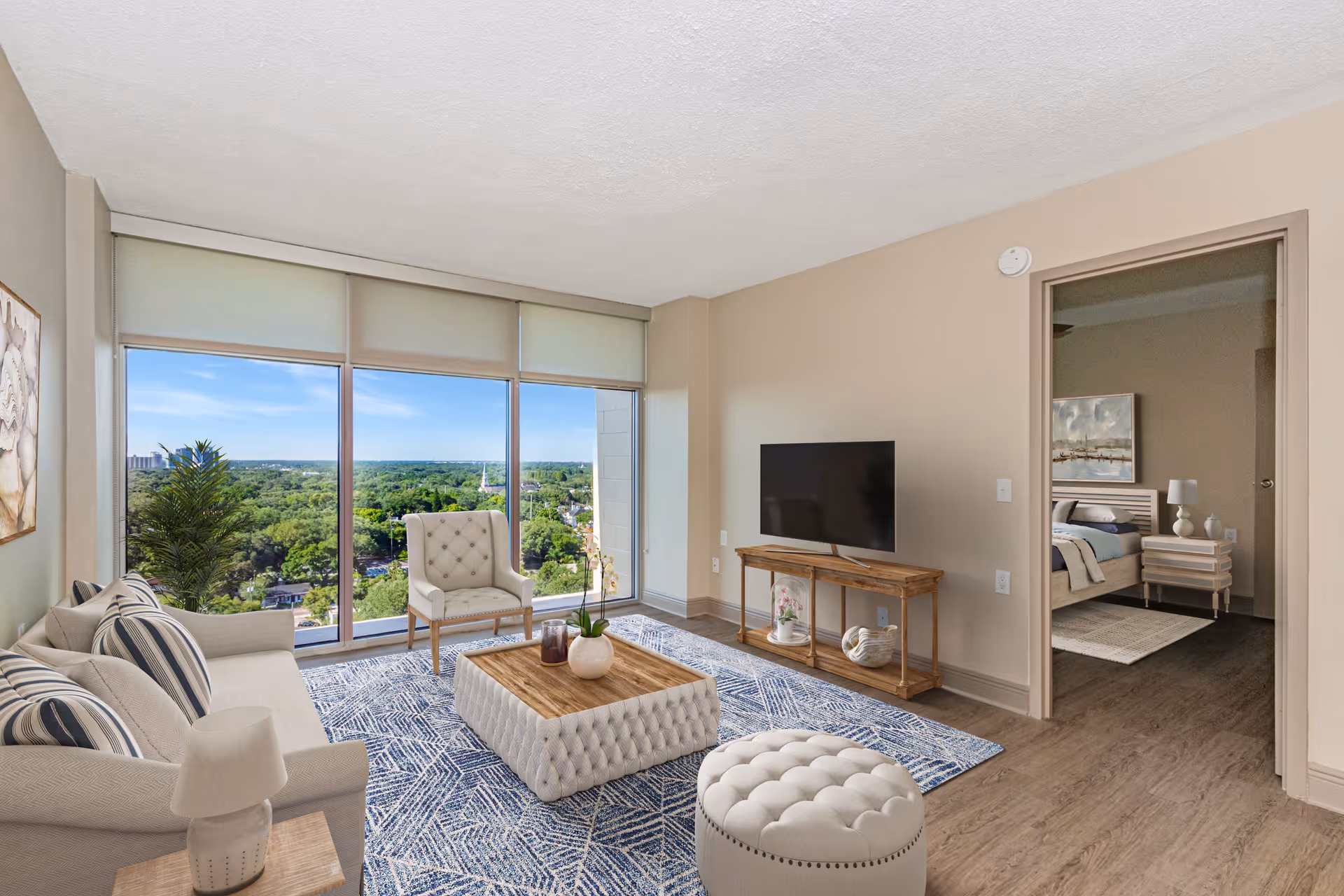 A bright living room with large floor-to-ceiling windows offering a view of green trees and a blue sky. The room features a beige sofa with striped pillows, a tufted ottoman coffee table with a wooden top, a beige armchair, a round tufted ottoman, a wooden TV stand with a flat-screen TV, and a blue patterned rug. A doorway leads to a bedroom with a bed, nightstand, lamp, and wall art.