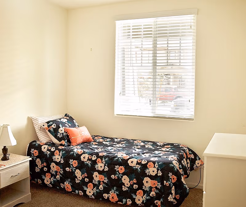 A small bedroom with a single bed covered in a dark floral bedspread. There are two pillows on the bed, one striped and one orange with the word 'love' on it. A white nightstand with a small lamp is next to the bed. A window with white blinds lets in natural light, and there is a white dresser on the right side of the room.