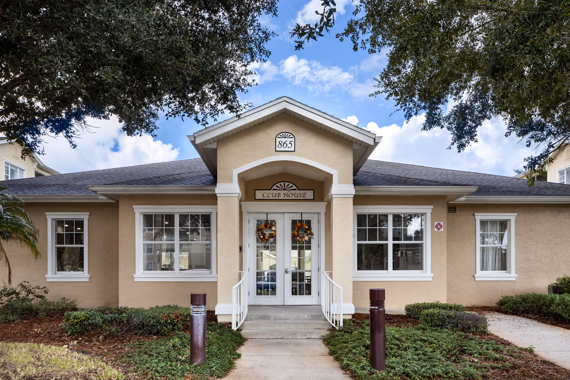 Exterior front view of a beige clubhouse building with white trim and a gray roof. The entrance has double glass doors decorated with wreaths and a small set of stairs with white railings. The building number 865 is displayed above the entrance, and there are windows on either side of the door. The scene is framed by trees and landscaping with bushes and mulch.
