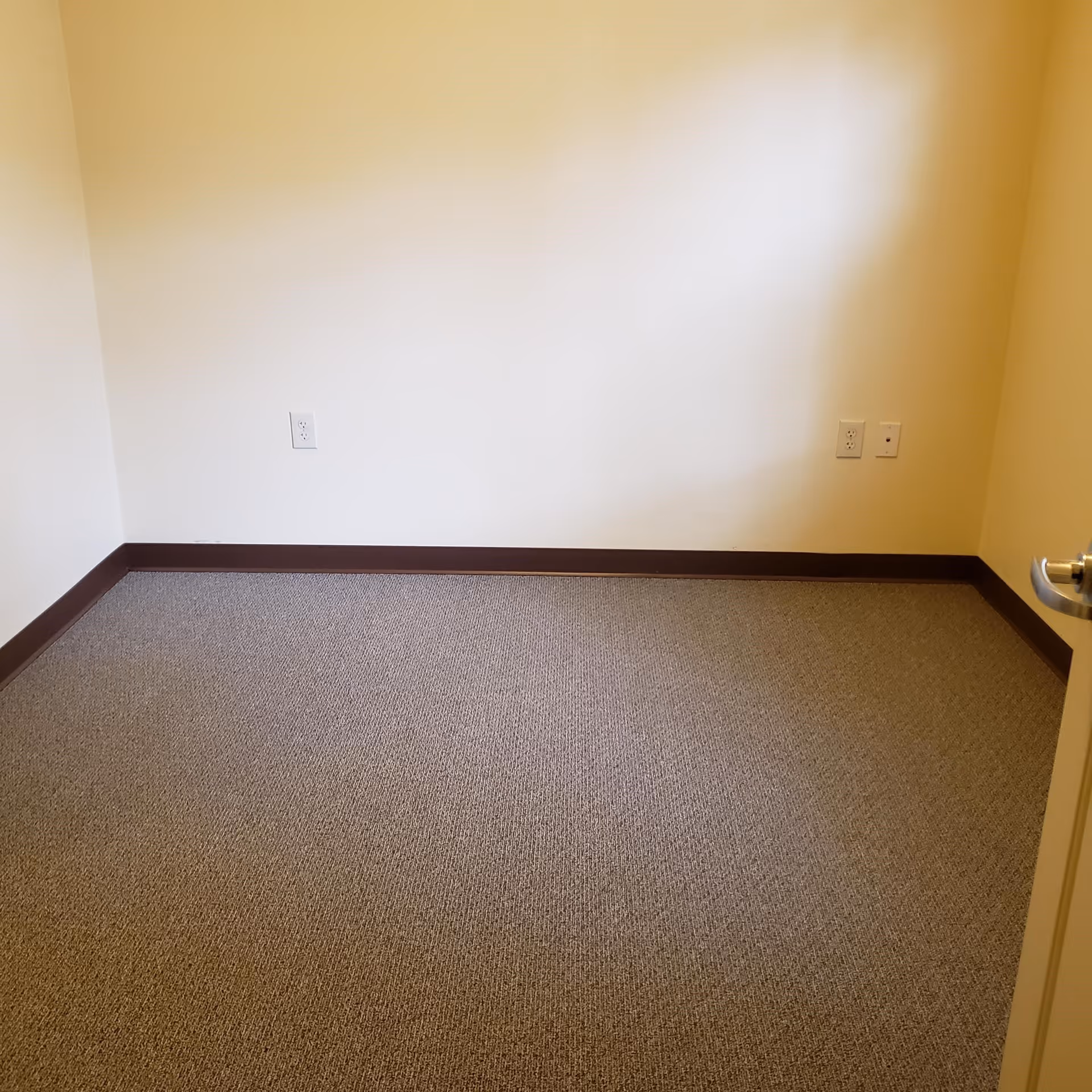 Empty room with beige walls, brown baseboards, and carpeted floor. There are three electrical outlets on the walls and a partially visible door with a silver handle on the right side.