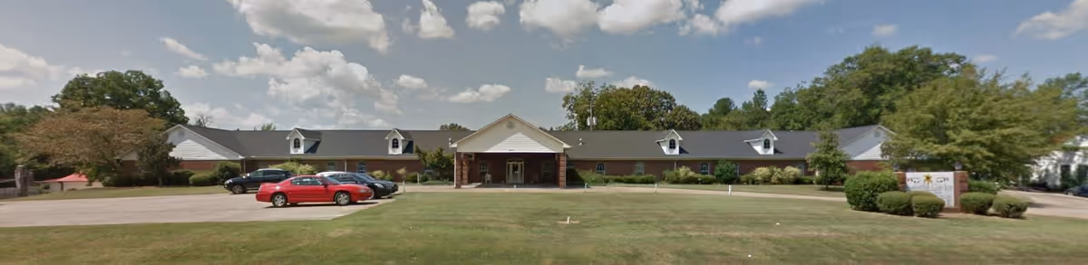 Front exterior view of Samaritan Garden facility showing a single-story building with a covered entrance, several parked cars in the driveway, and a grassy lawn with trees and shrubs under a partly cloudy sky.