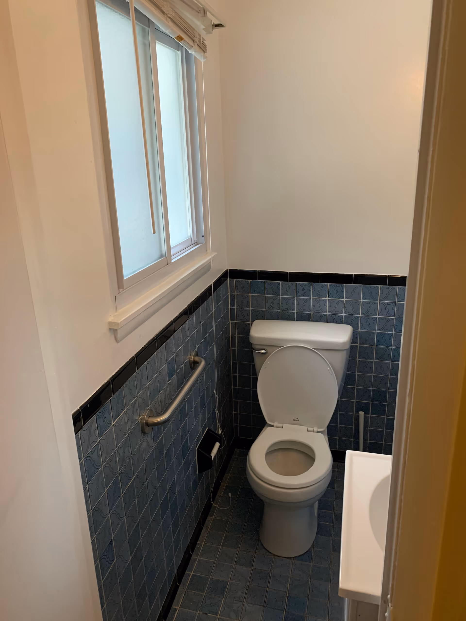 Small bathroom with a white toilet, blue tiled walls with a black border, a metal grab bar on the left wall, a toilet paper holder, a window with blinds above the grab bar, and a partial view of a white sink on the right.