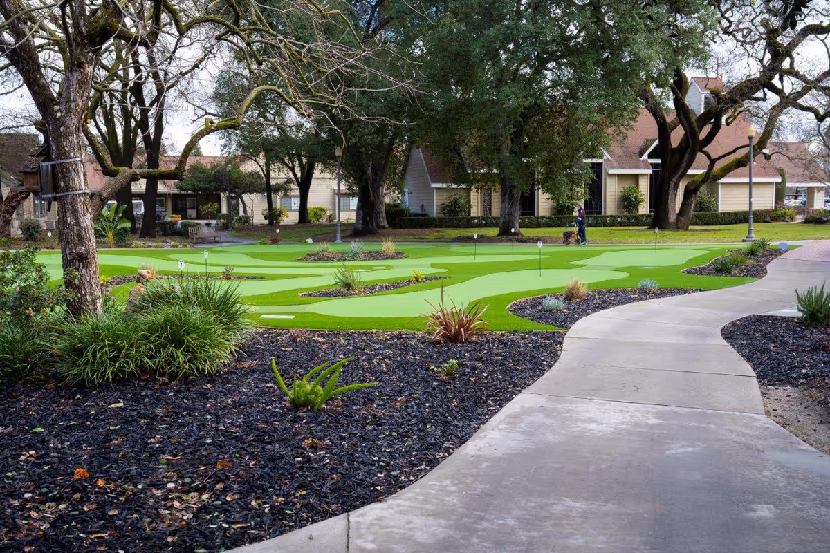 A landscaped outdoor area at O'Connor Woods featuring a putting green with several small flags, surrounded by trees, shrubs, and a curved concrete pathway. Residential-style buildings are visible in the background, and a person is walking with a walker near the putting green.