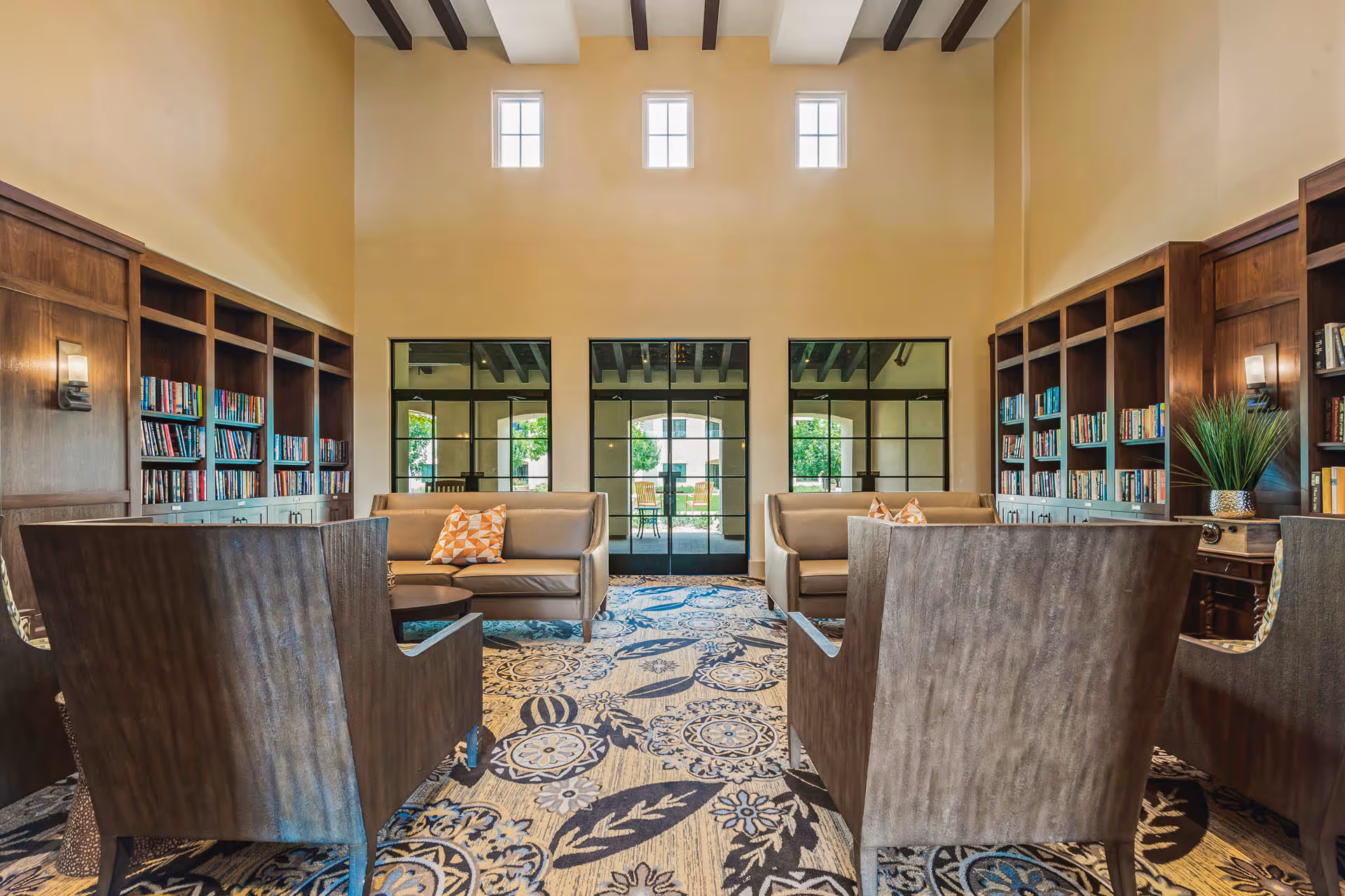A spacious communal lounge with built-in bookshelves, sofas and armchairs under a tall ceiling with small high windows and glass doors to a patio.